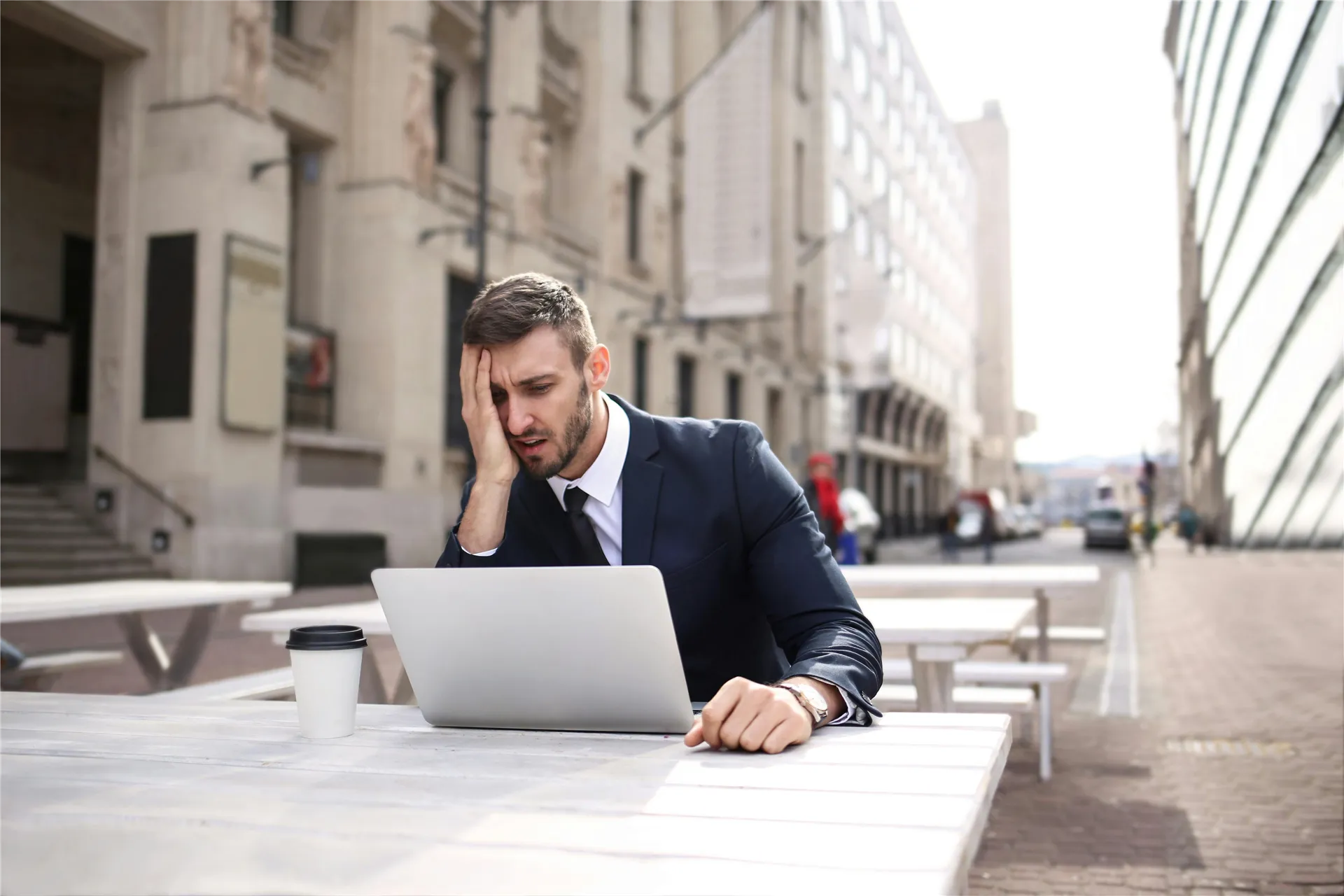 A businessman in a black suit looks stressed while working outdoors on a laptop with coffee.